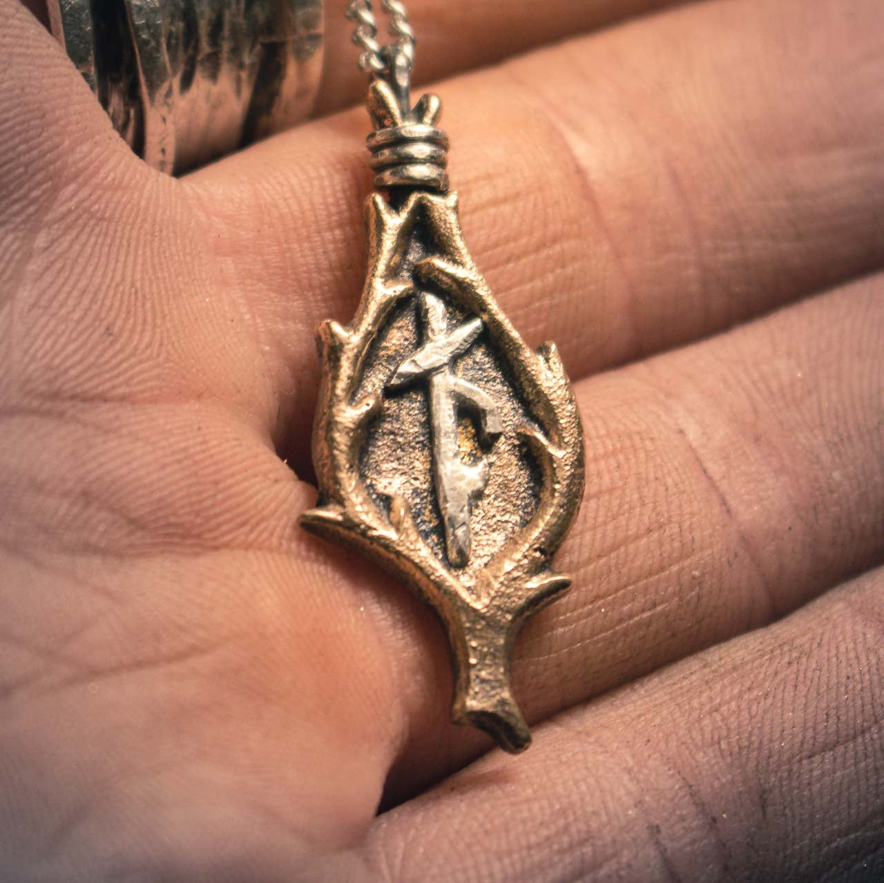 Close-up macro shot of a bronze Viking amulet with combined runes, suspended on a silver chain against a black surface
